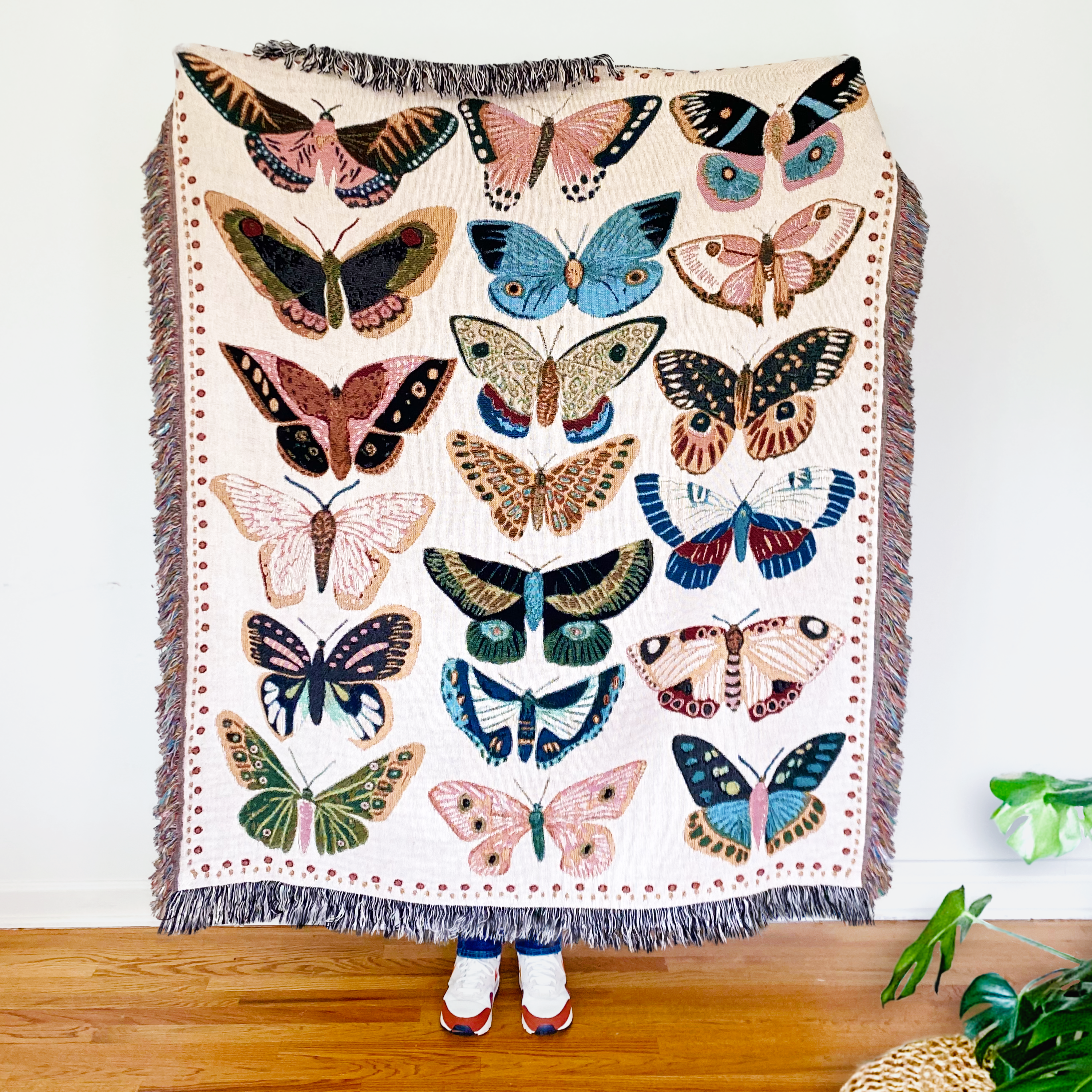 Woven throw blanket featuring vibrant hand-illustrated butterflies in pink, turquoise, yellow, coral, sage green, and rust orange on soft cream background. Monarchs, swallowtails, painted ladies, and blue morpho butterflies create joyful nature-inspired celebration with decorative fringe border.