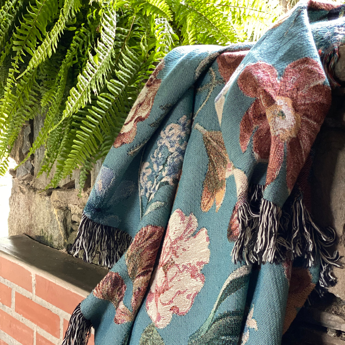 The botanical blanket draped over a brick ledge beside a fern, a peach bloom in soft folds. Nostalgic and gentle.