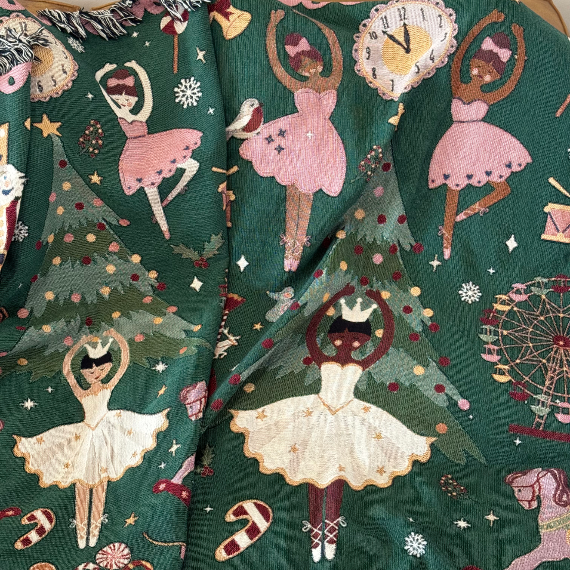 Close-up of the Nutcracker woven blanket on deep green, showing all five prima ballerinas together: two in white star-dotted tutus with little crowns and three in pink dresses, their skin tones ranging from fair to deep brown. They dance among Christmas trees, a clock at midnight, candy canes, a robin, a toy drum and a Ferris wheel. Festive and joyful.