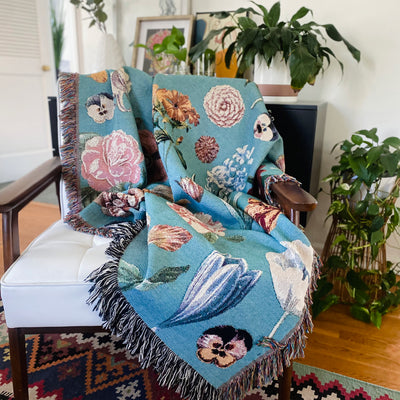 The botanical blanket draped over a white chair beside a plant shelf, its blooms soft on blue. Romantic and calm.